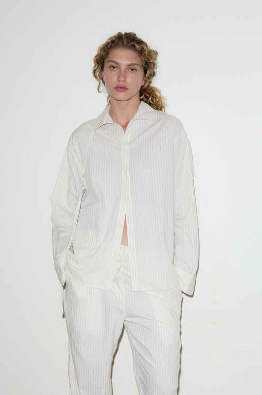 A person with curly hair stands against a white wall, wearing the Lapel Bow Shirt—a loose-fitting, white pinstripe button-up—with matching pants and hands in their pockets.
