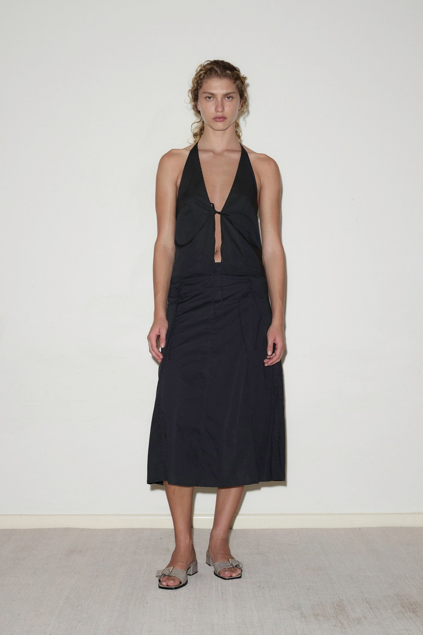 A woman stands against a white wall, wearing the Double Lace Up Skirt in black with a matching sleeveless halter top, her curly hair tied back and paired with heeled sandals.
