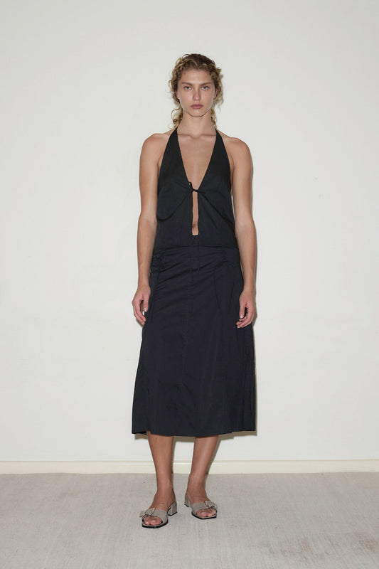 A woman with light brown, wavy hair pulled back stands against a white wall, wearing a sleeveless black halter top and the Double Lace Up Skirt. She completes her look with gray open-toe sandals.