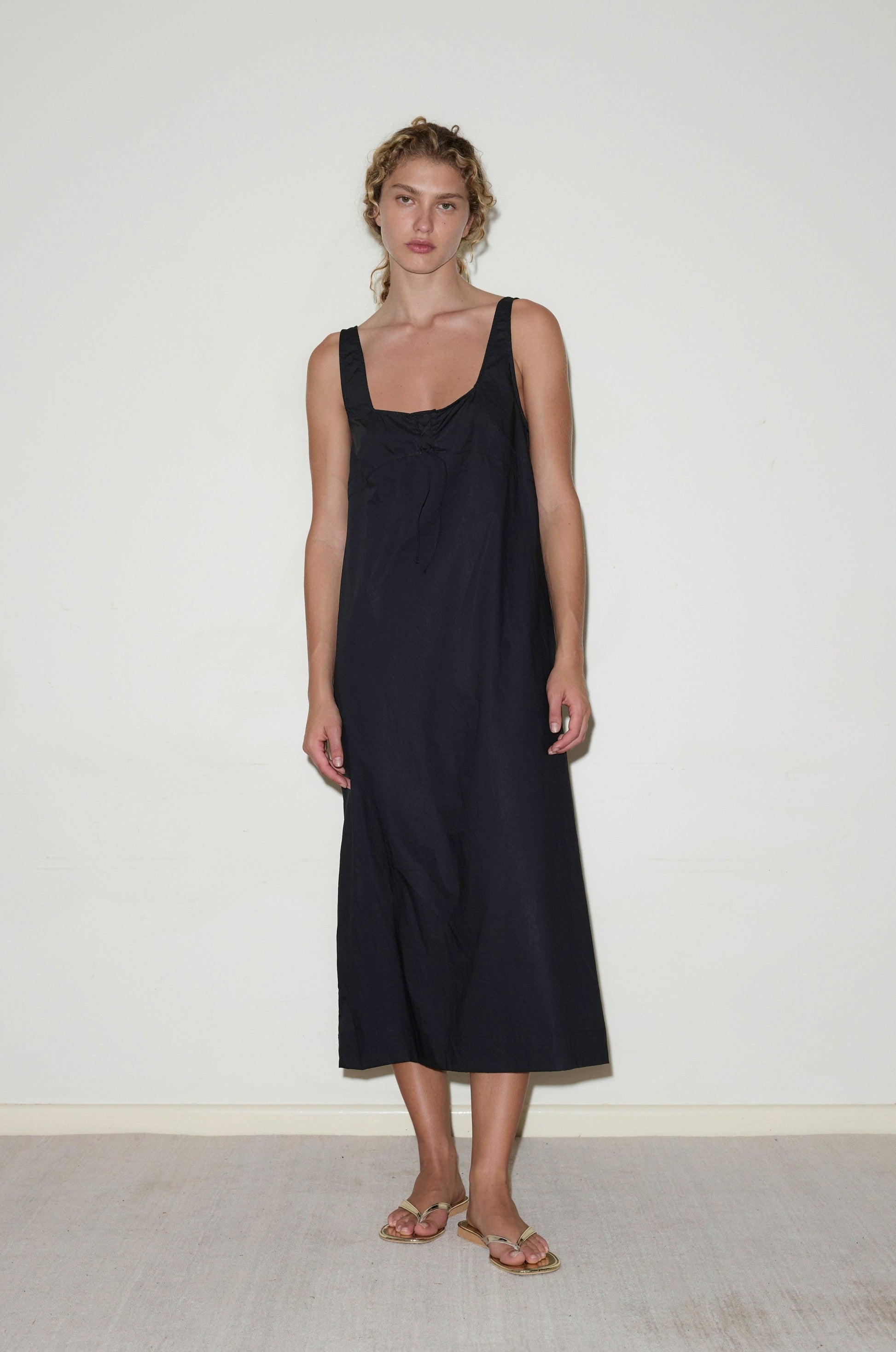 A woman with light brown, wavy hair tied back stands indoors against a plain white wall, wearing the Eyelet Dress - Black from Deiji Studios and beige sandals. She has a neutral expression.