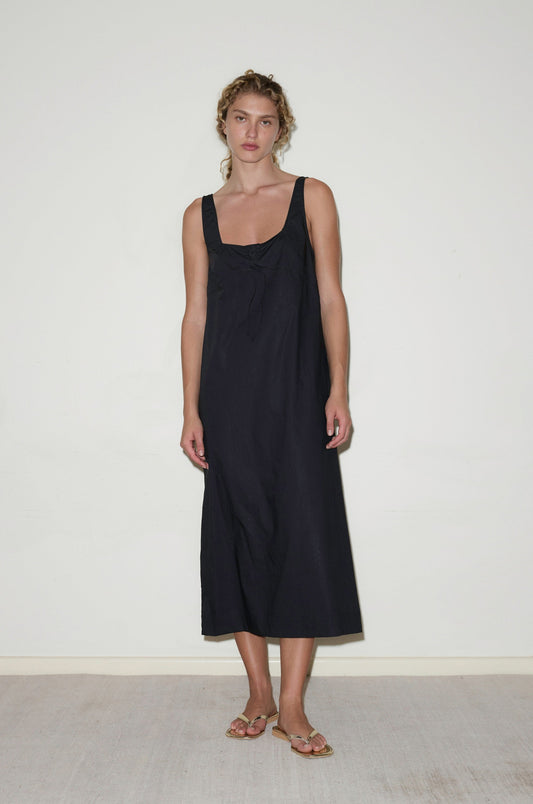 A woman with light brown, wavy hair tied back stands indoors against a plain white wall, wearing the Eyelet Dress - Black from Deiji Studios and beige sandals. She has a neutral expression.