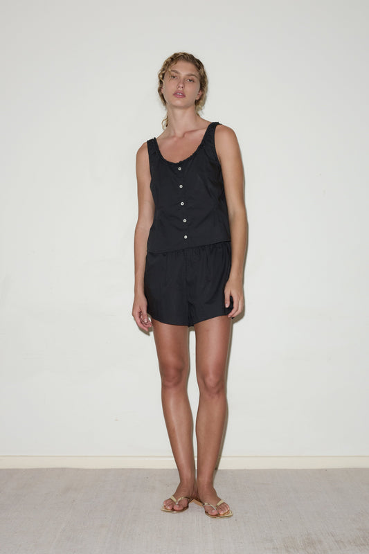 A person stands against a plain white wall, wearing the PJ Short Set—a sleeveless black top with buttons and matching black shorts. They have loose, wavy light brown hair and are wearing flat sandals.