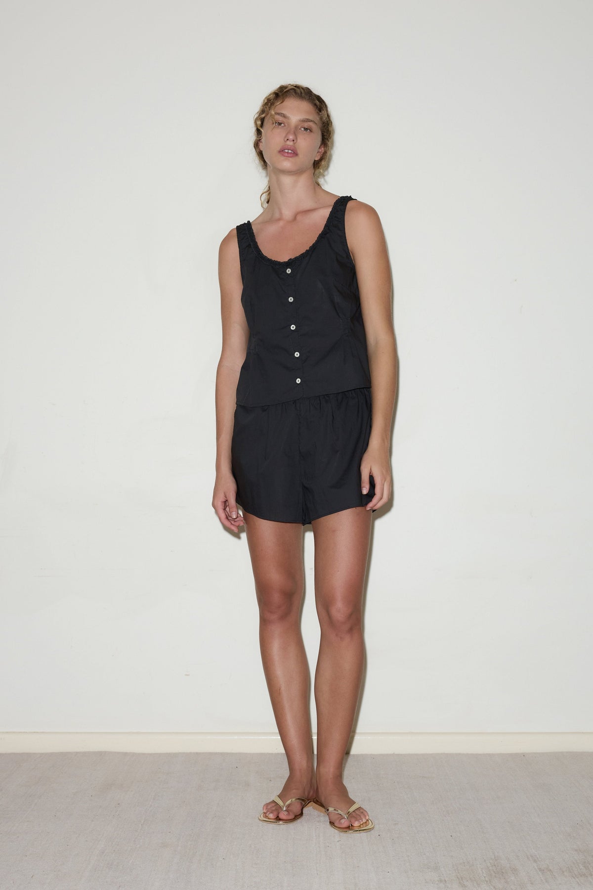A woman with wavy hair and a neutral expression stands indoors against a white wall, wearing the PJ Short Set—a sleeveless black buttoned top and matching shorts. She pairs the minimal look with sandals.