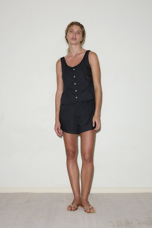 A woman with wavy hair and a neutral expression stands indoors against a white wall, wearing the PJ Short Set—a sleeveless black buttoned top and matching shorts. She pairs the minimal look with sandals.