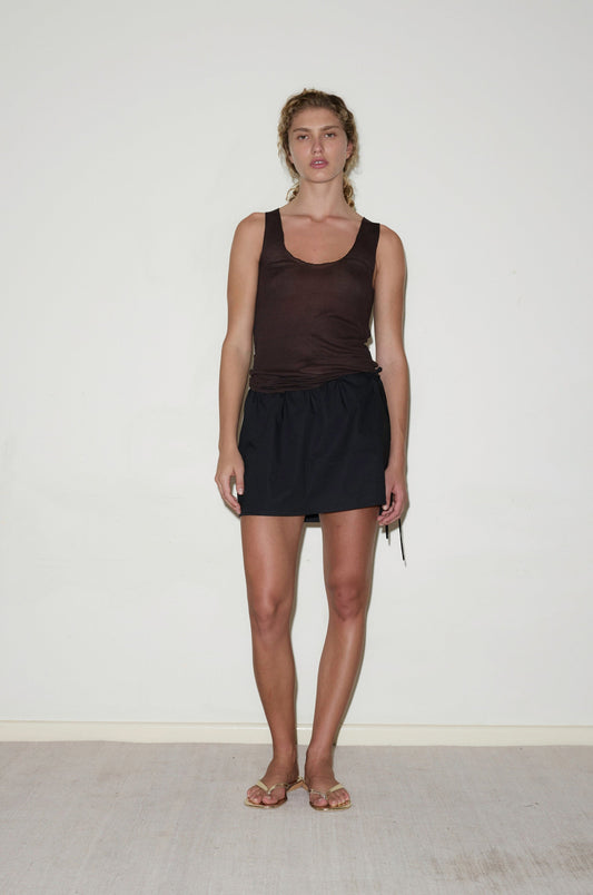 A woman with curly hair pulled back stands against a white wall, wearing the Block Mini Skirt with a sleeveless brown top and flat sandals. She looks directly at the camera, her expression neutral.