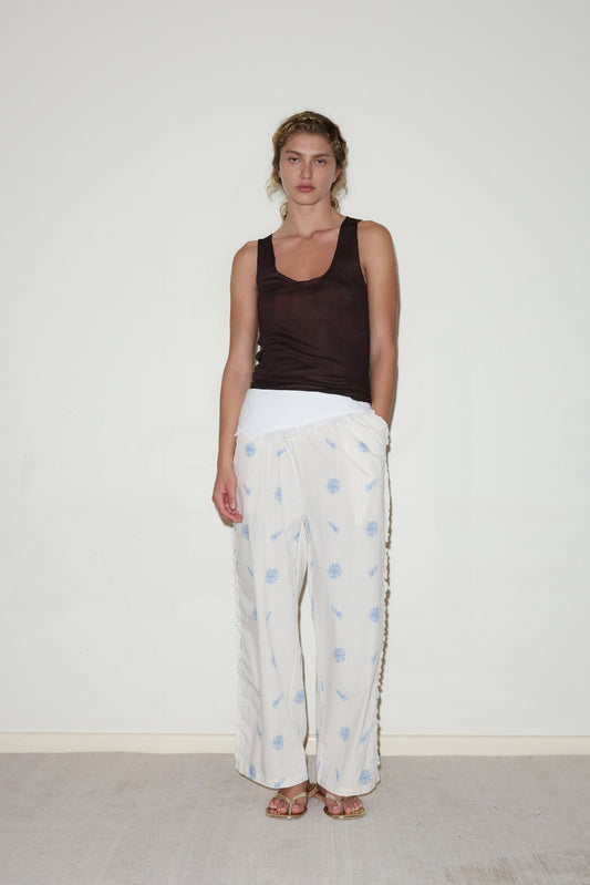 A person stands against a white wall wearing the Ruffle Pant in a white and blue pattern, paired with a dark sleeveless top and sandals. Their light brown hair is pulled back, and they have one hand in their pocket.