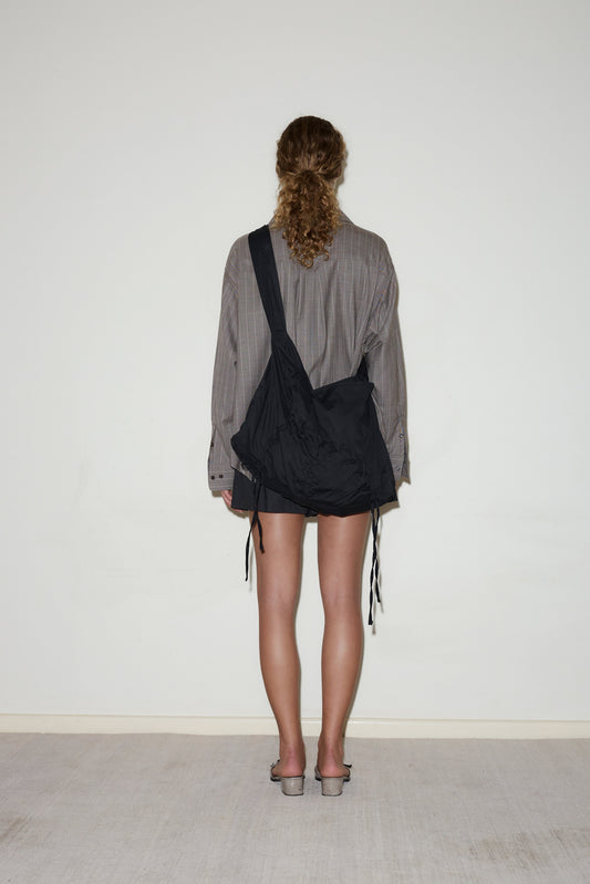 A person with curly hair faces a white wall indoors on a light carpet, wearing a loose long-sleeved shirt, black shorts, slide sandals, and carrying the Crossed Tie Bag—a large black shoulder bag.