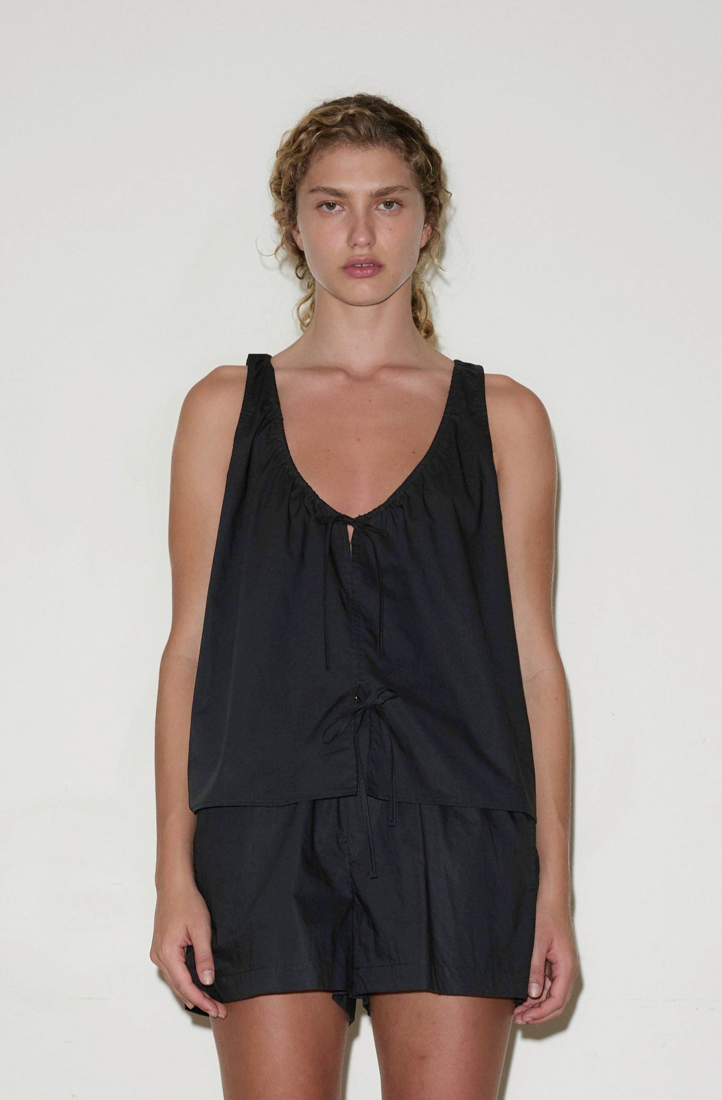 A woman with curly hair wears the Cotton Bind Top - Black by Deiji Studios and matching shorts, standing against a plain white background and looking directly at the camera with a neutral expression.