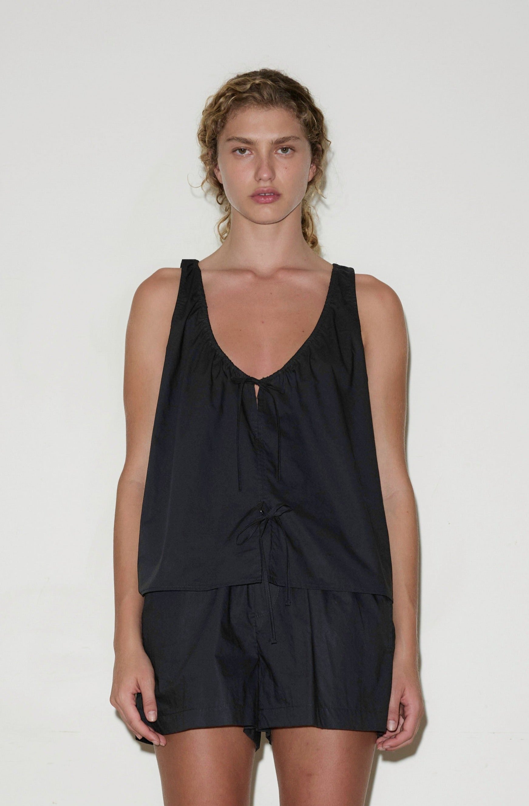 A woman with curly hair wears the Cotton Bind Top - Black by Deiji Studios and matching shorts, standing against a plain white background and looking directly at the camera with a neutral expression.