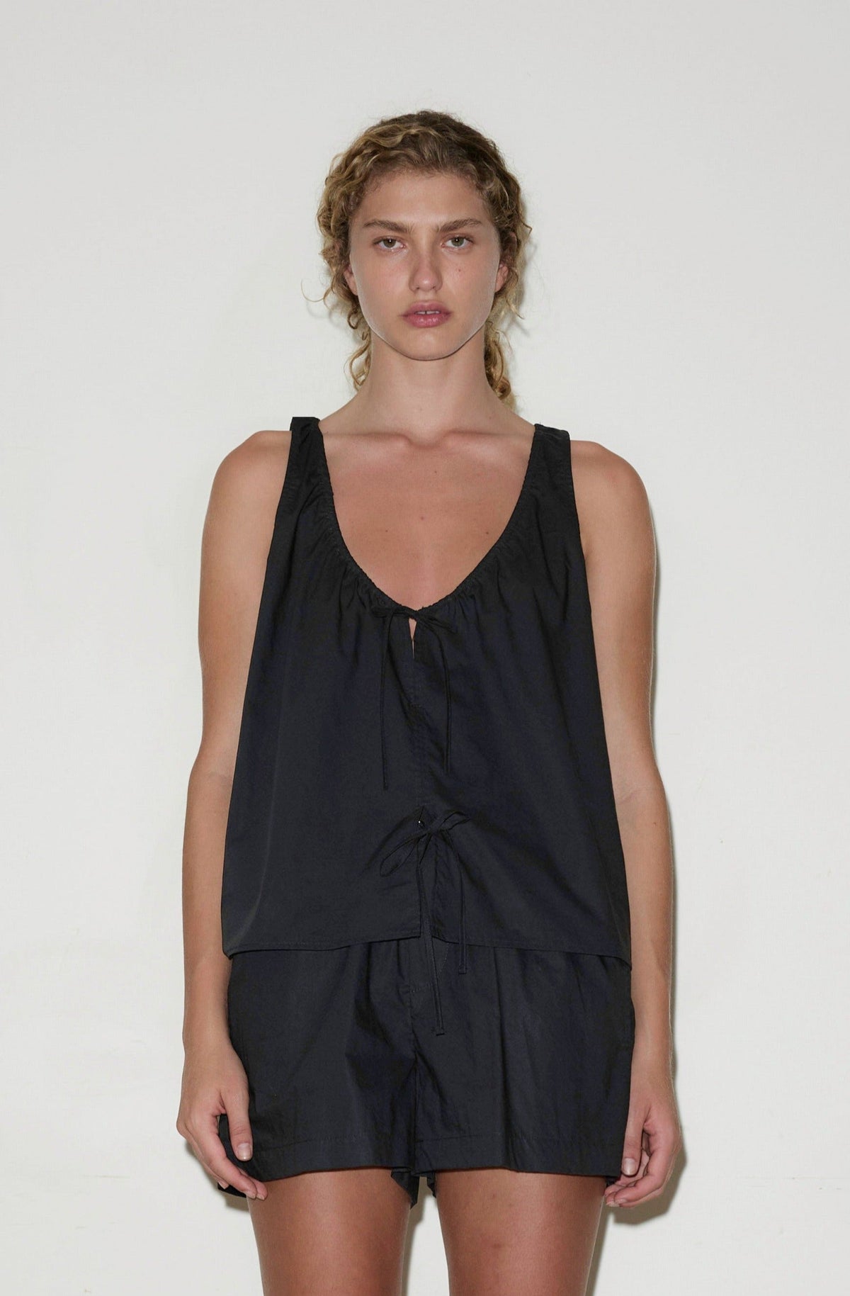 A woman with curly hair wears the Cotton Bind Top - Black by Deiji Studios and matching shorts, standing against a plain white background and looking directly at the camera with a neutral expression.