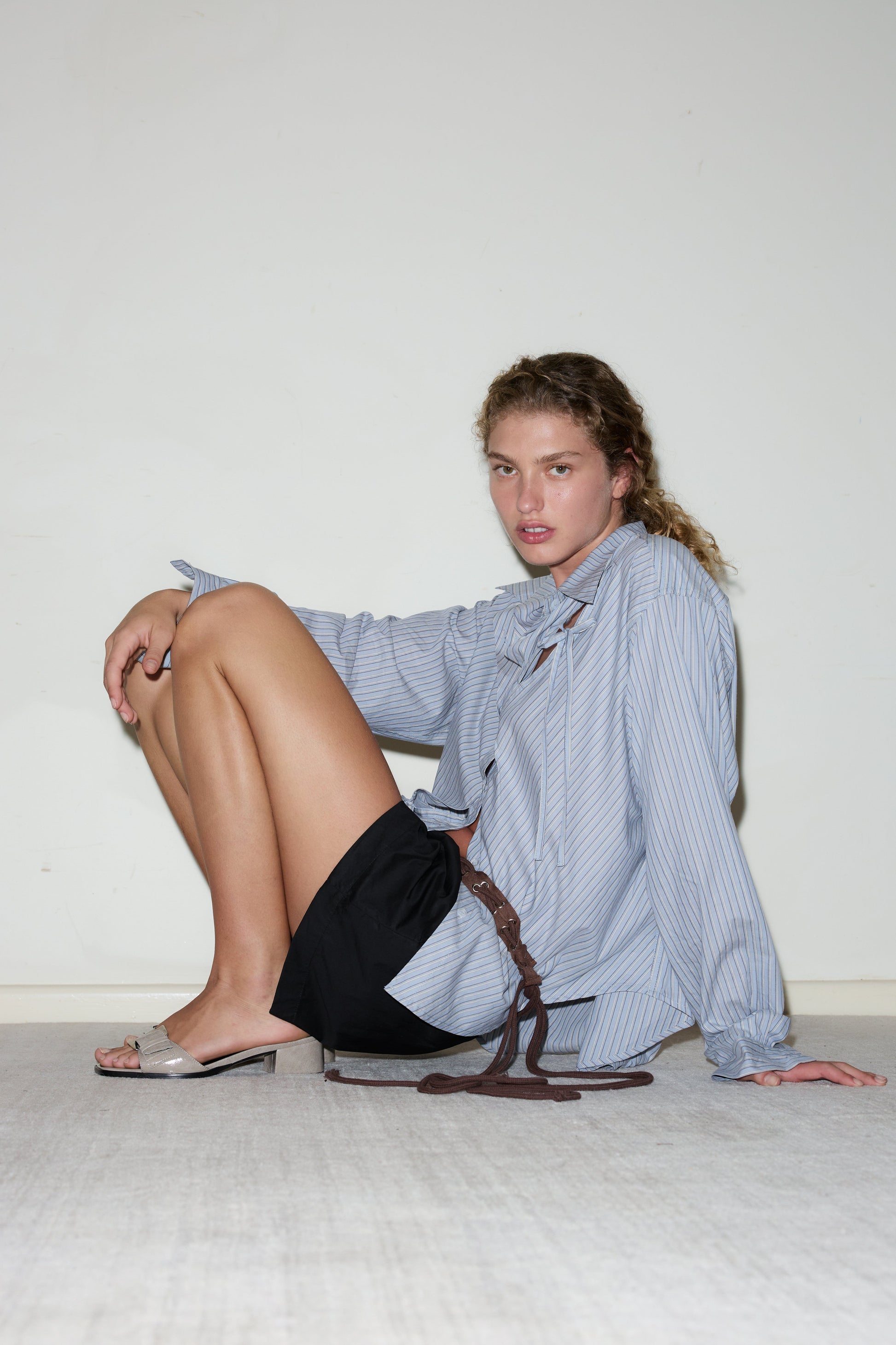 A person with curly hair sits on the floor against a plain wall, wearing a loose striped shirt, black shorts with a brown Eyelet Belt, and open-toed heels, looking at the camera with a neutral expression.