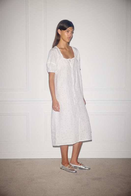 A woman stands indoors against a white paneled wall, wearing the Curved Seam Midi Dress—a loose, white, midi-length dress with short puff sleeves and small dotted patterns—paired with silver ballet flats.