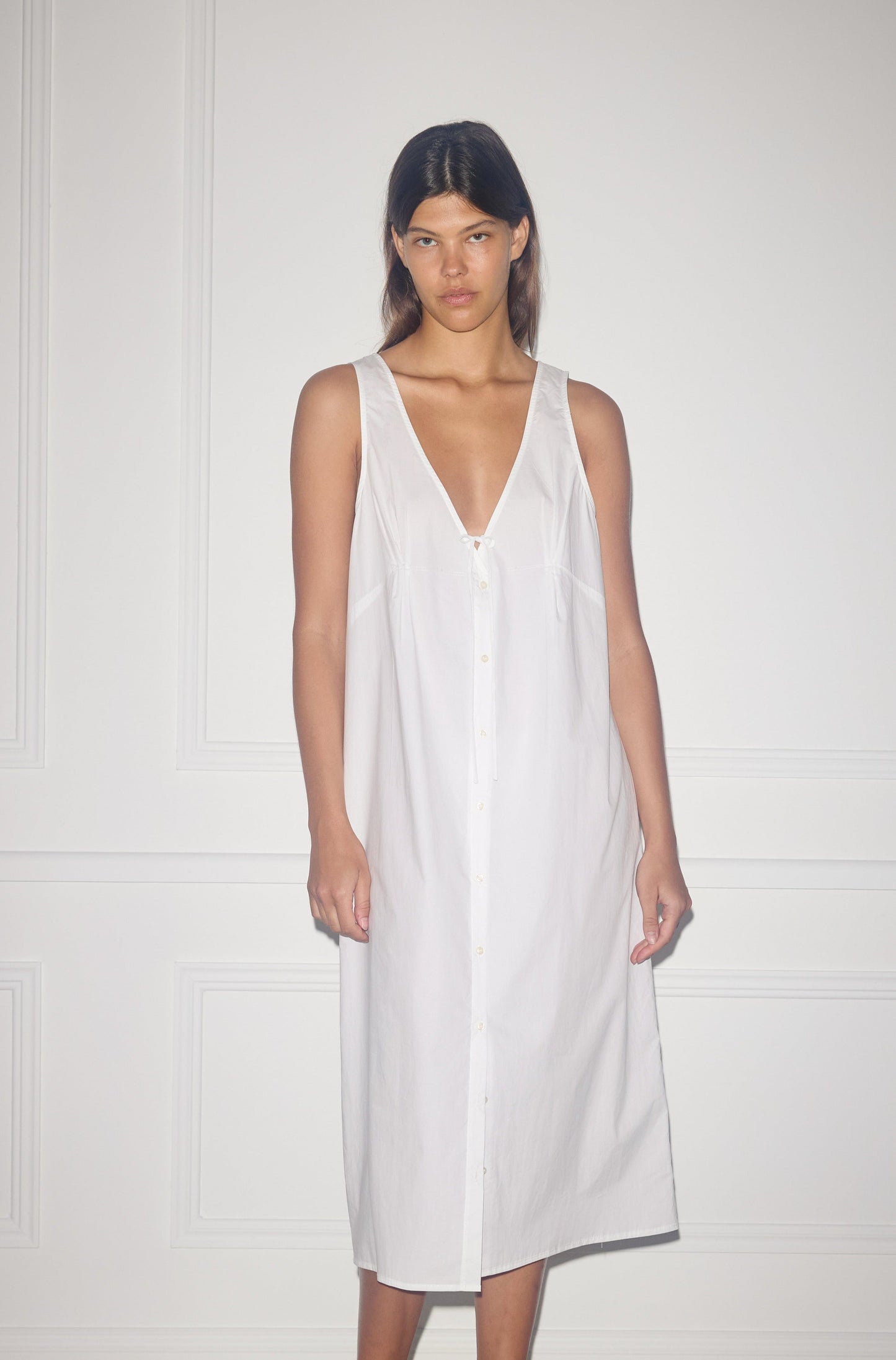 A person stands against a white wall wearing the Tuck Tie Dress—a sleeveless, loose-fitting white button-down that falls below the knees with a deep V-neck and relaxed, minimalist style.