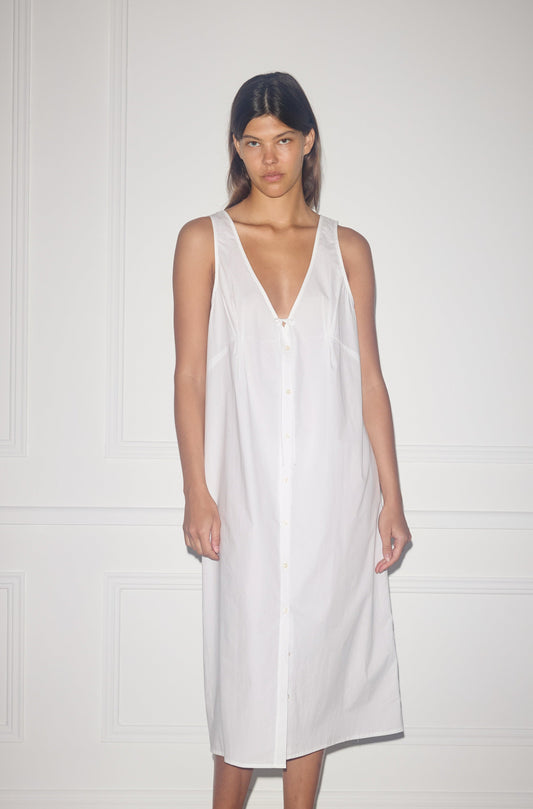 A person stands against a white wall wearing the Tuck Tie Dress—a sleeveless, loose-fitting white button-down that falls below the knees with a deep V-neck and relaxed, minimalist style.