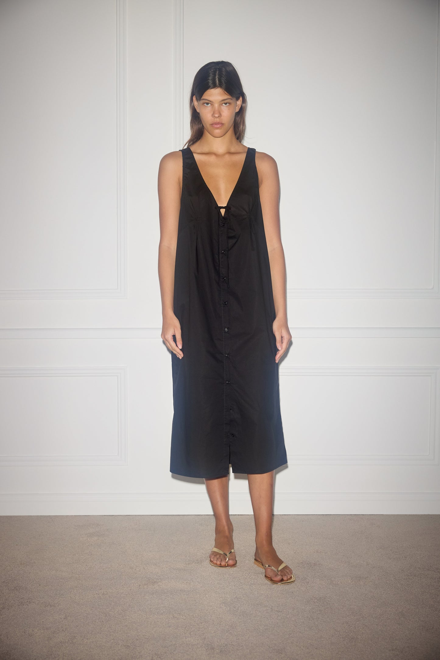 A woman with straight dark hair and a neutral expression stands against a white wall, wearing the sleeveless, knee-length black Tuck Tie Dress with front buttons, paired with beige sandals.