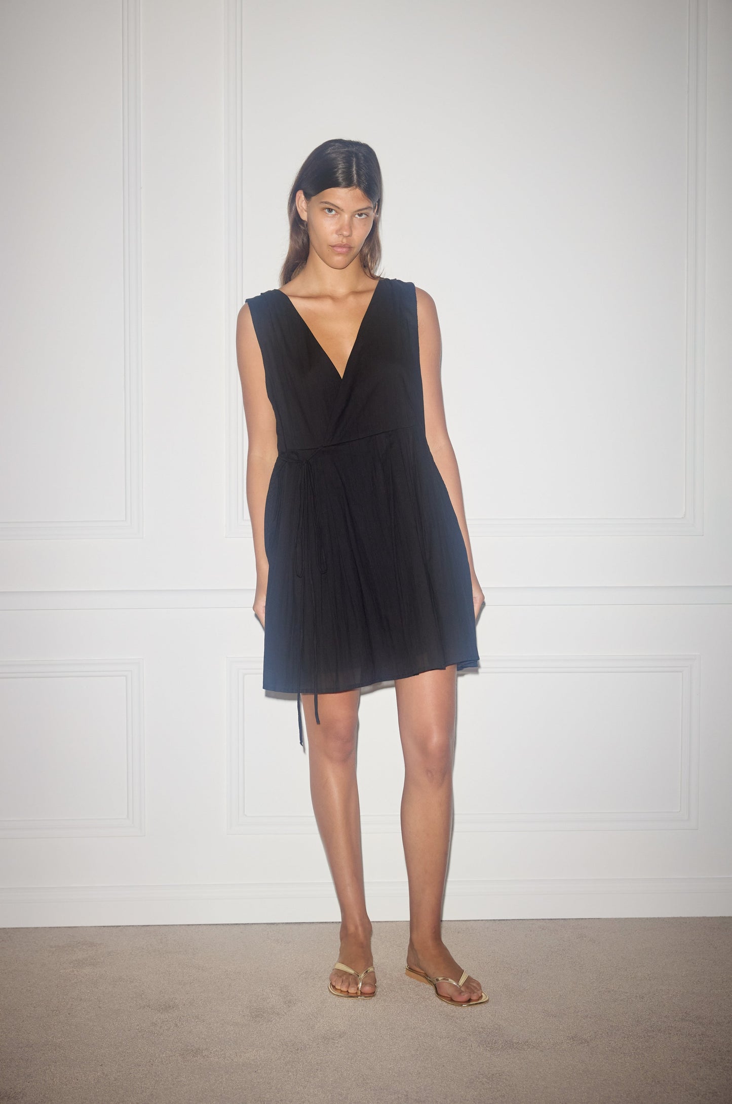 A woman with straight dark hair stands against a white paneled wall, wearing the sleeveless black Wrap Tie Dress and gold sandals. She has a neutral expression and relaxed arms by her sides.