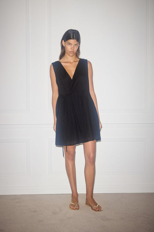 A woman with straight dark hair stands against a white paneled wall, wearing the sleeveless black Wrap Tie Dress and gold sandals. She has a neutral expression and relaxed arms by her sides.