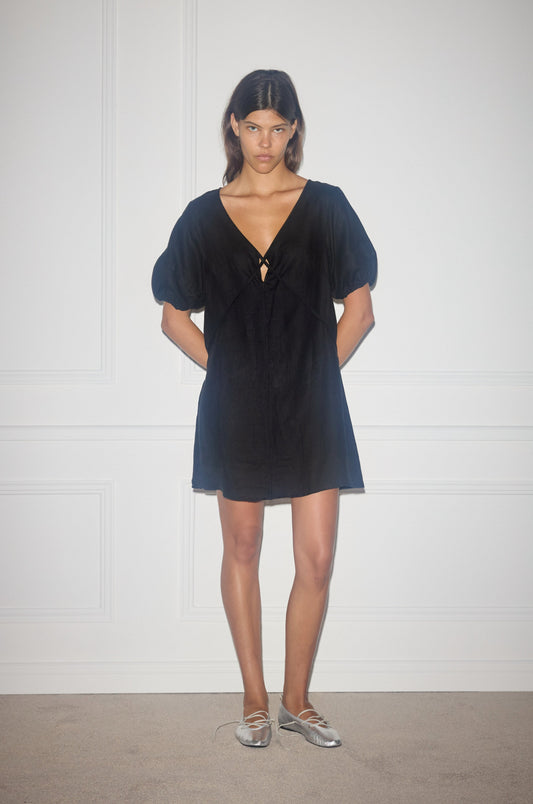 A woman stands indoors against a white paneled wall, wearing the Tie Seamed Short Dress—a short black dress with a deep V-neck and puffed sleeves—paired with silver ballet flats. Her hands are behind her back and she has a neutral expression.
