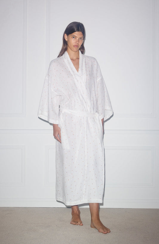 A person stands indoors against a white wall, barefoot and wearing the 02 Robe—a long white robe with a subtle dotted pattern and belted waist. They have a neutral expression.