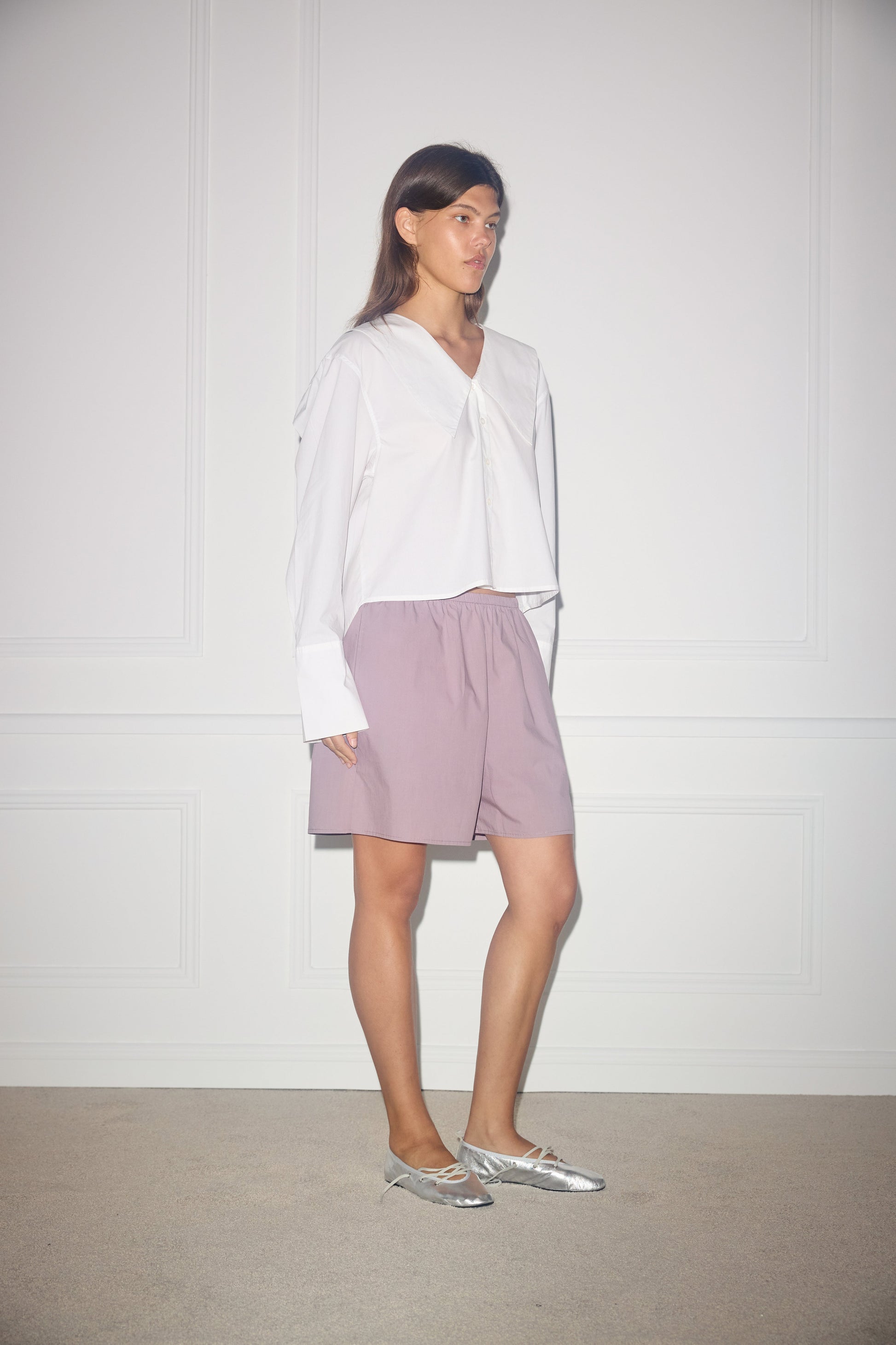 A woman with straight brown hair and a neutral expression stands indoors against a white wall, wearing a loose white blouse, silver flats, and light purple knee-length mid shorts.