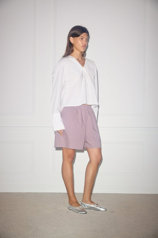 A woman with straight brown hair and a neutral expression stands indoors against a white wall, wearing a loose white blouse, silver flats, and light purple knee-length mid shorts.