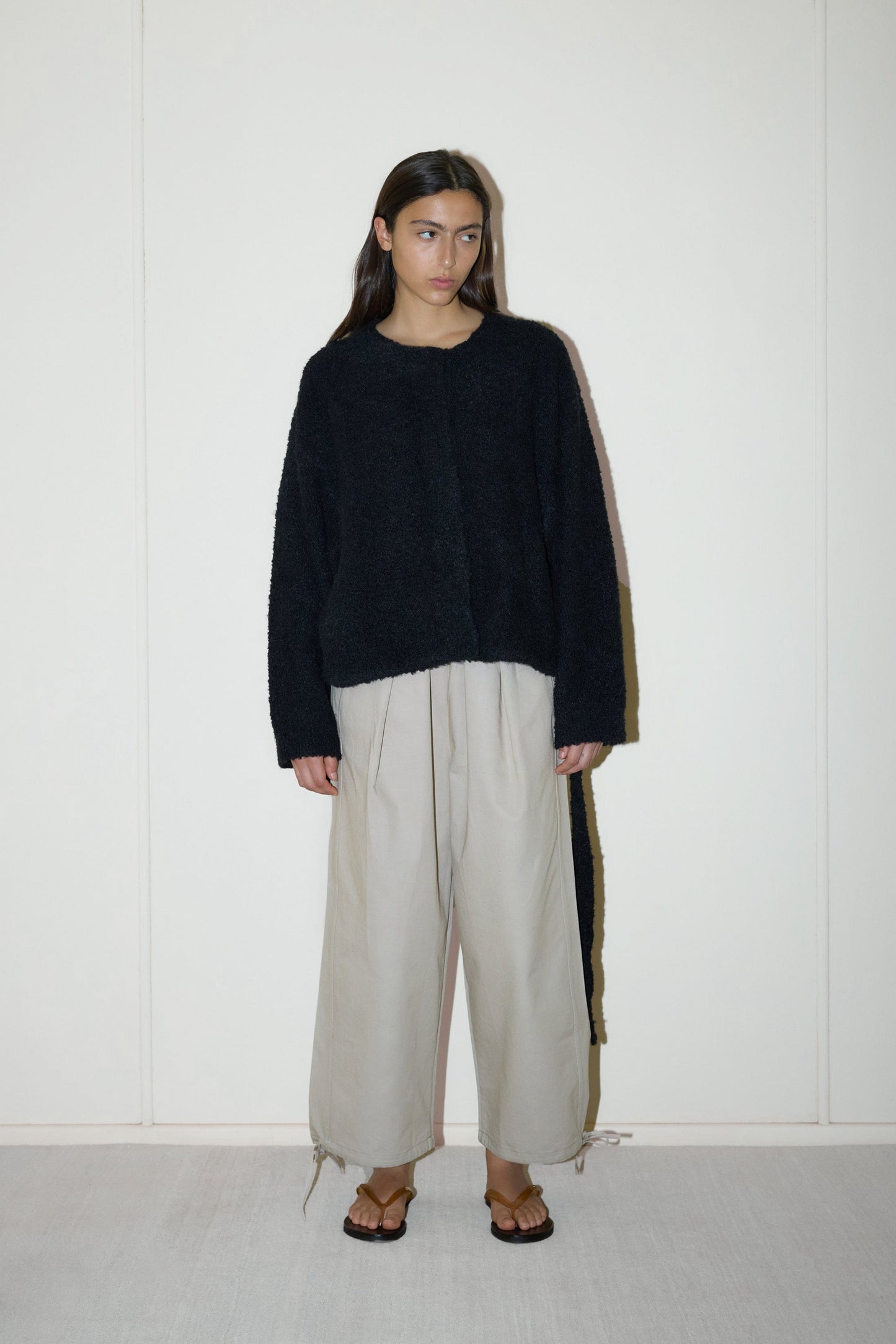darked haired female model wears a black relaxed and textured knit features a round shaped neckline with press stud front closure, removable self tie belt with long sleeves and high back neckline, paired with cream structured cotton twill trouser featuring a drawcord hem and waist and deep side pockets designed to sit mid-low waist for a relaxed look.