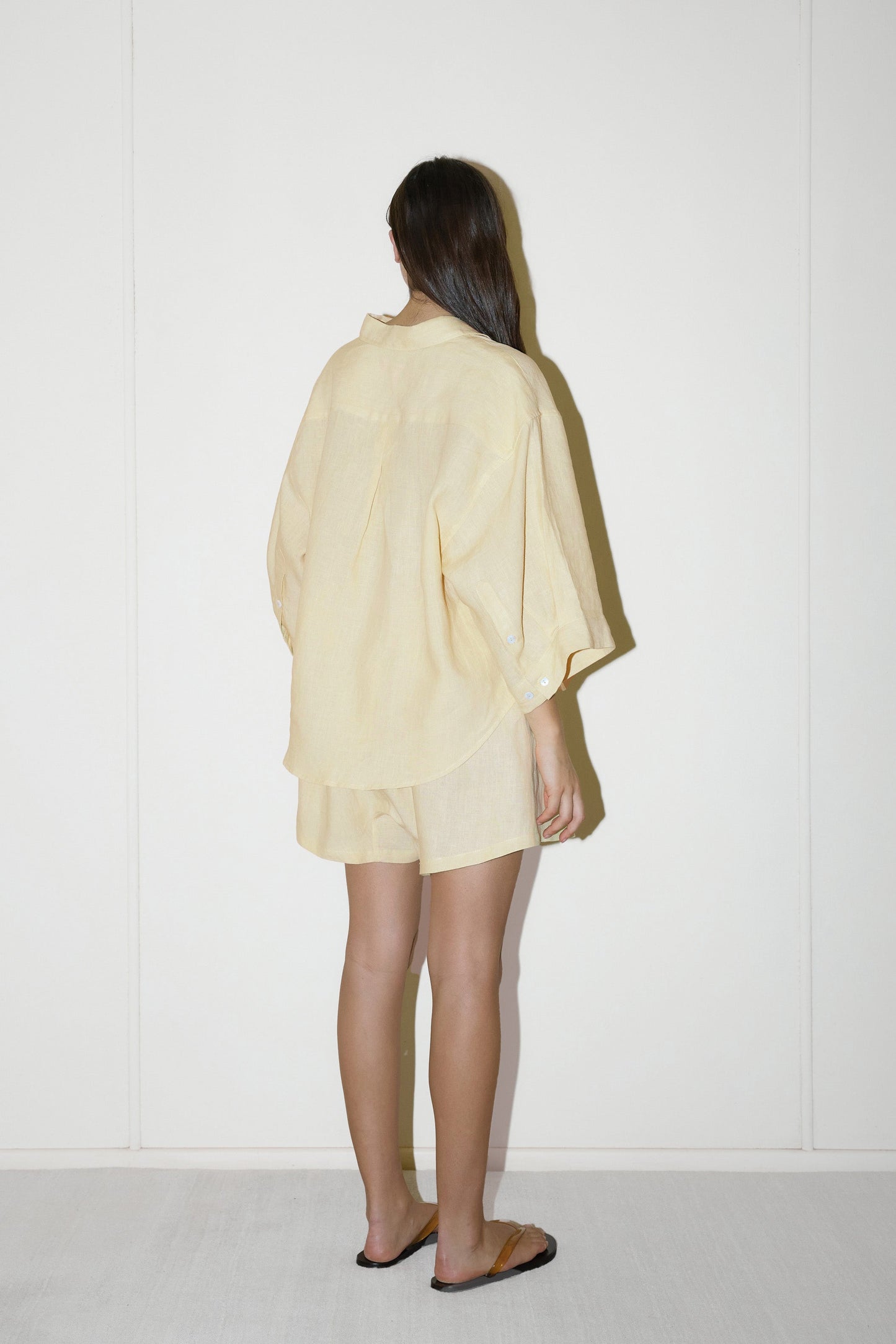 dark haired female model wears a butter yellow shirt and short set in an oversized box fit linen shirt with wide arms and a front pocket designed to sit mid rise, the loose fitting boxer style shorts have a faux button down fly and an elastic waist for extra comfort