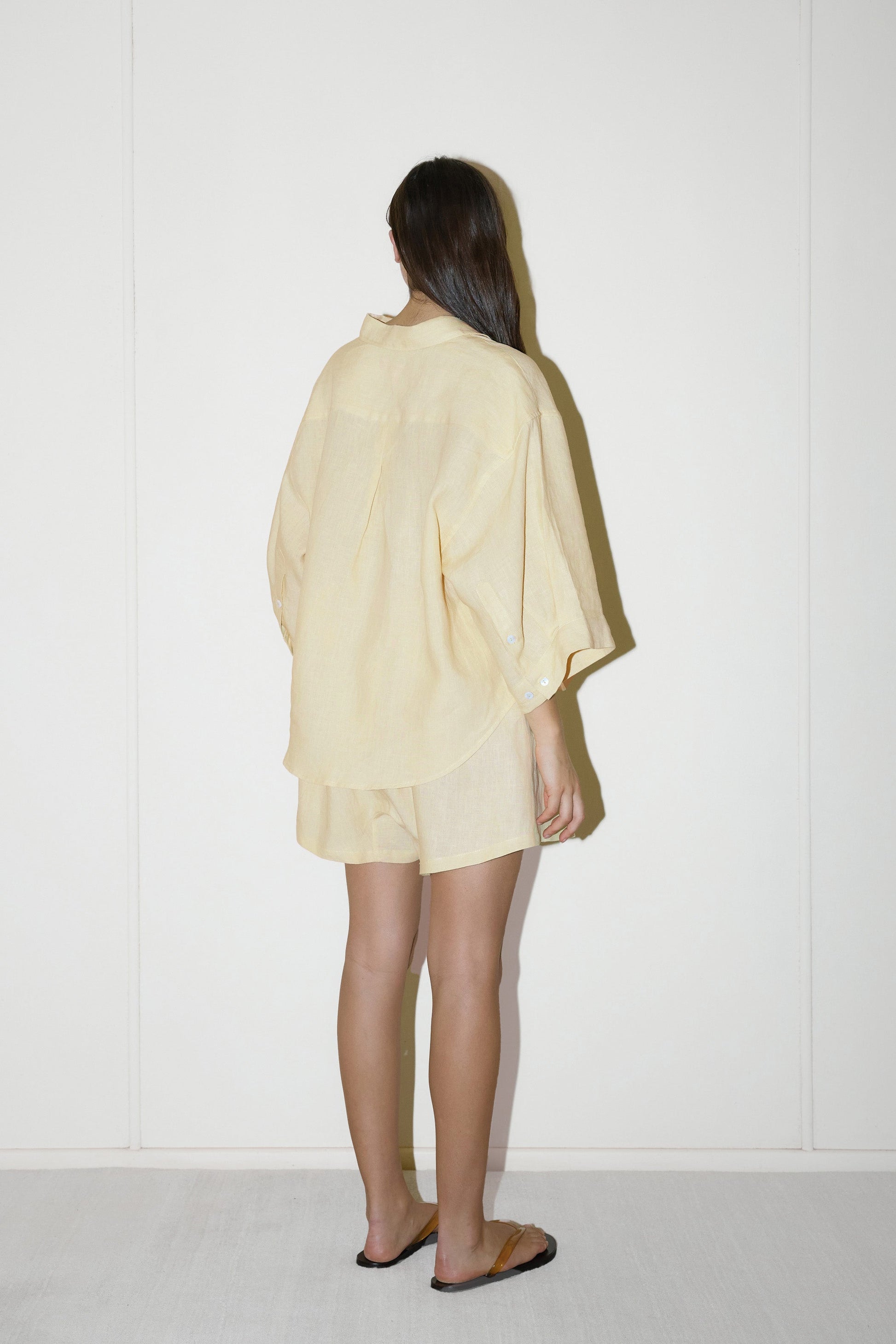 dark haired female model wears a butter yellow shirt and short set in an oversized box fit linen shirt with wide arms and a front pocket designed to sit mid rise, the loose fitting boxer style shorts have a faux button down fly and an elastic waist for extra comfort