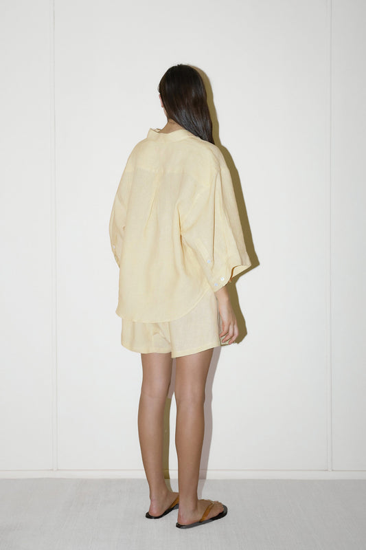 dark haired female model wears a butter yellow shirt and short set in an oversized box fit linen shirt with wide arms and a front pocket designed to sit mid rise, the loose fitting boxer style shorts have a faux button down fly and an elastic waist for extra comfort