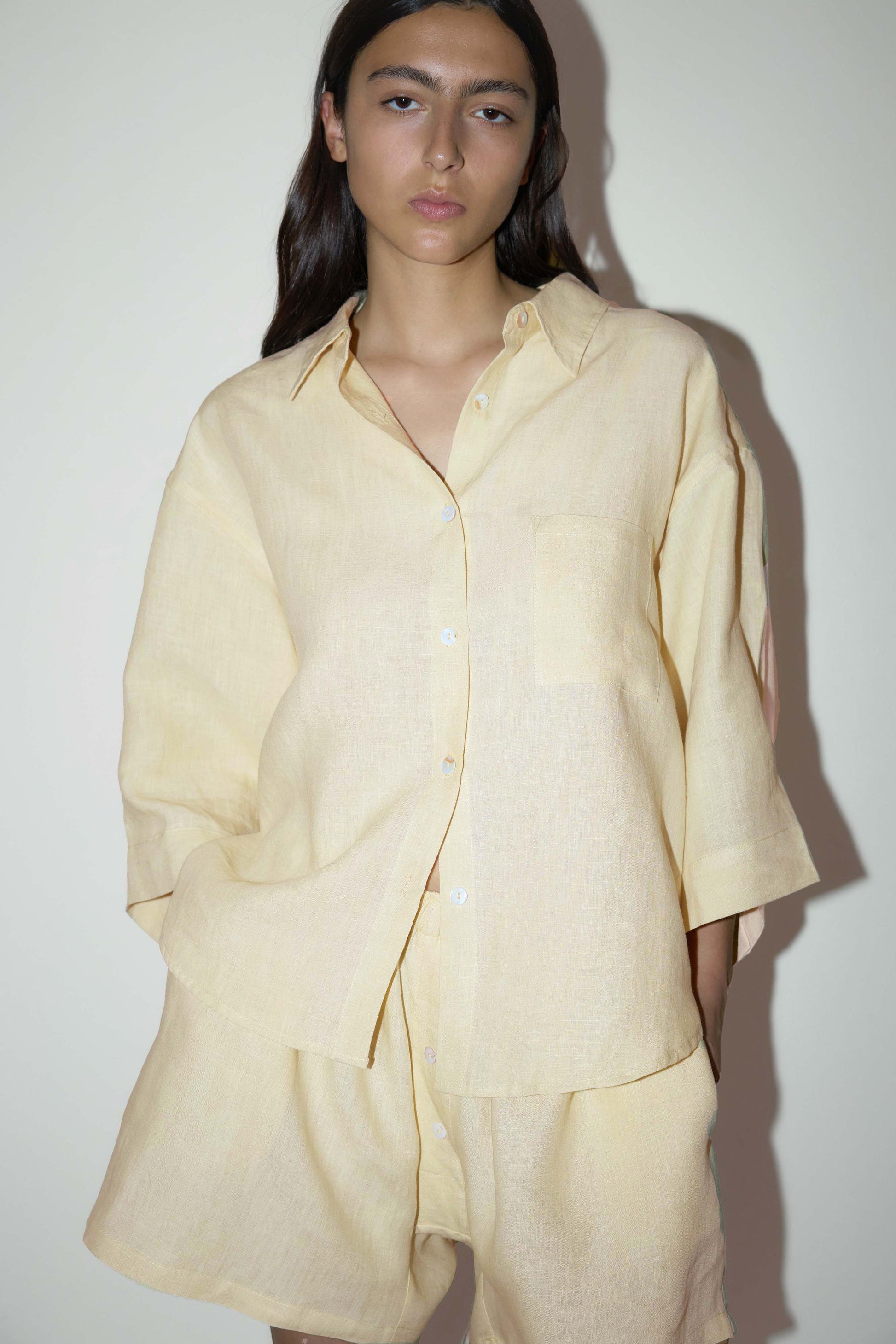 dark haired female model wears a butter yellow shirt and short set in an oversized box fit linen shirt with wide arms and a front pocket designed to sit mid rise, the loose fitting boxer style shorts have a faux button down fly and an elastic waist for extra comfort