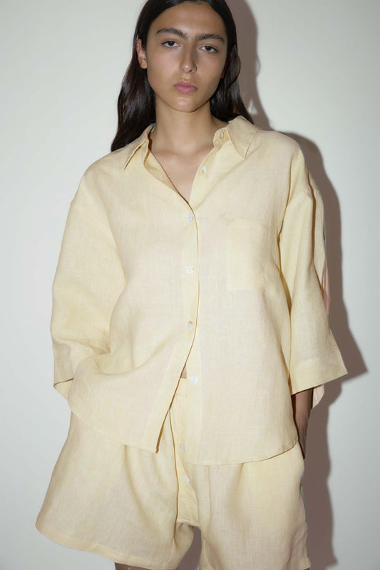 dark haired female model wears a butter yellow shirt and short set in an oversized box fit linen shirt with wide arms and a front pocket designed to sit mid rise, the loose fitting boxer style shorts have a faux button down fly and an elastic waist for extra comfort