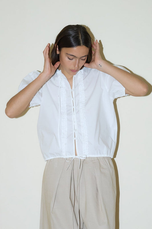 A person with straight brown hair looks down, hands near their head, wearing the Double Frill Top - White by Deiji Studios and light beige high-waisted pants against a plain light background.