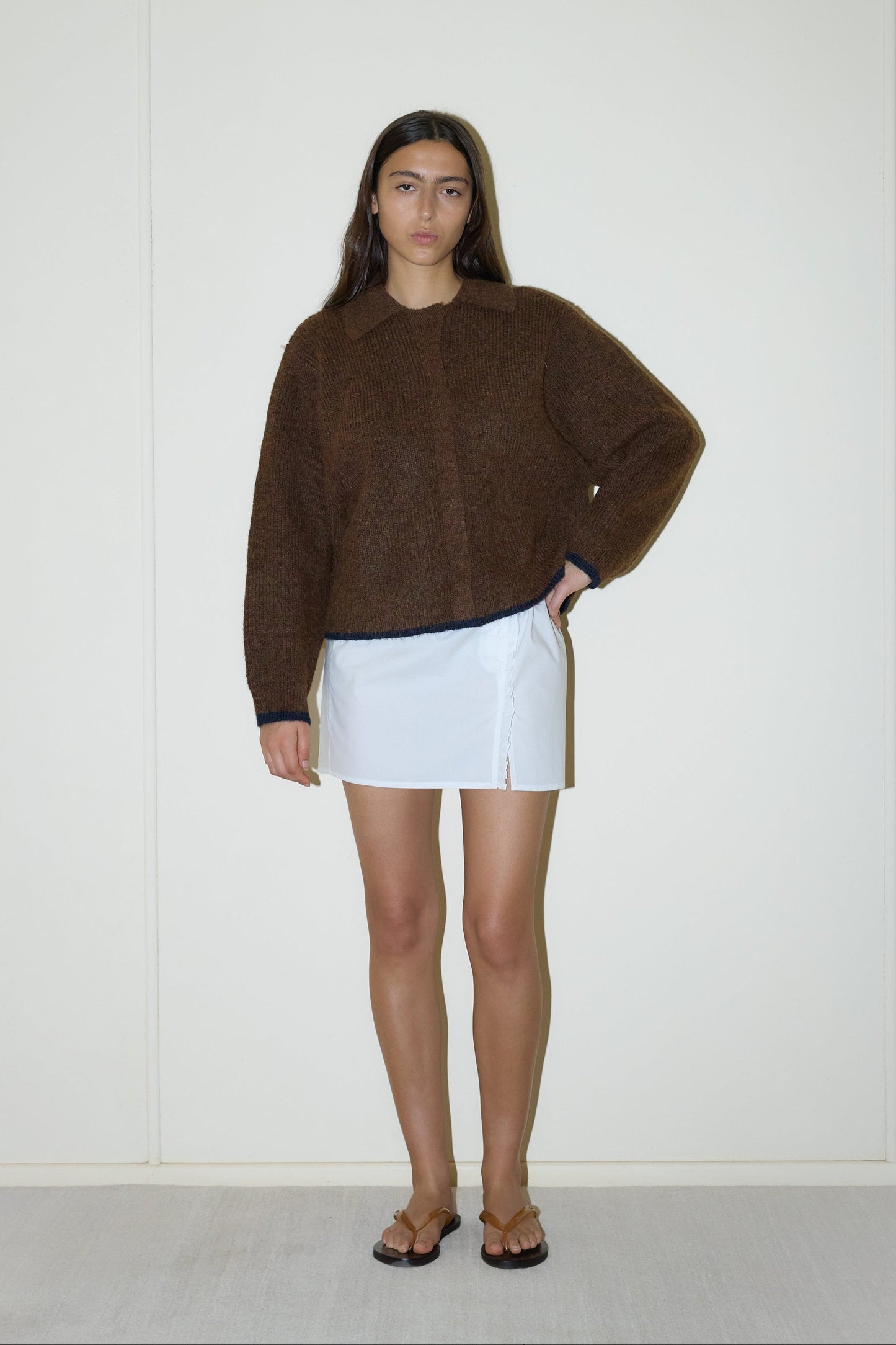 dark haired female model wears a brown elevated in a luxurious softly weighted and subtly lustrous sustainable knit blend featuring invisible press stud closure front, a relaxed collar and dropped shoulder, a gently curved arm for soft shaping and oversized fit for layering, paired with a white simple mini skirt designed to sit at low waist features adjustable tie and ruffle edge on side with button detail
