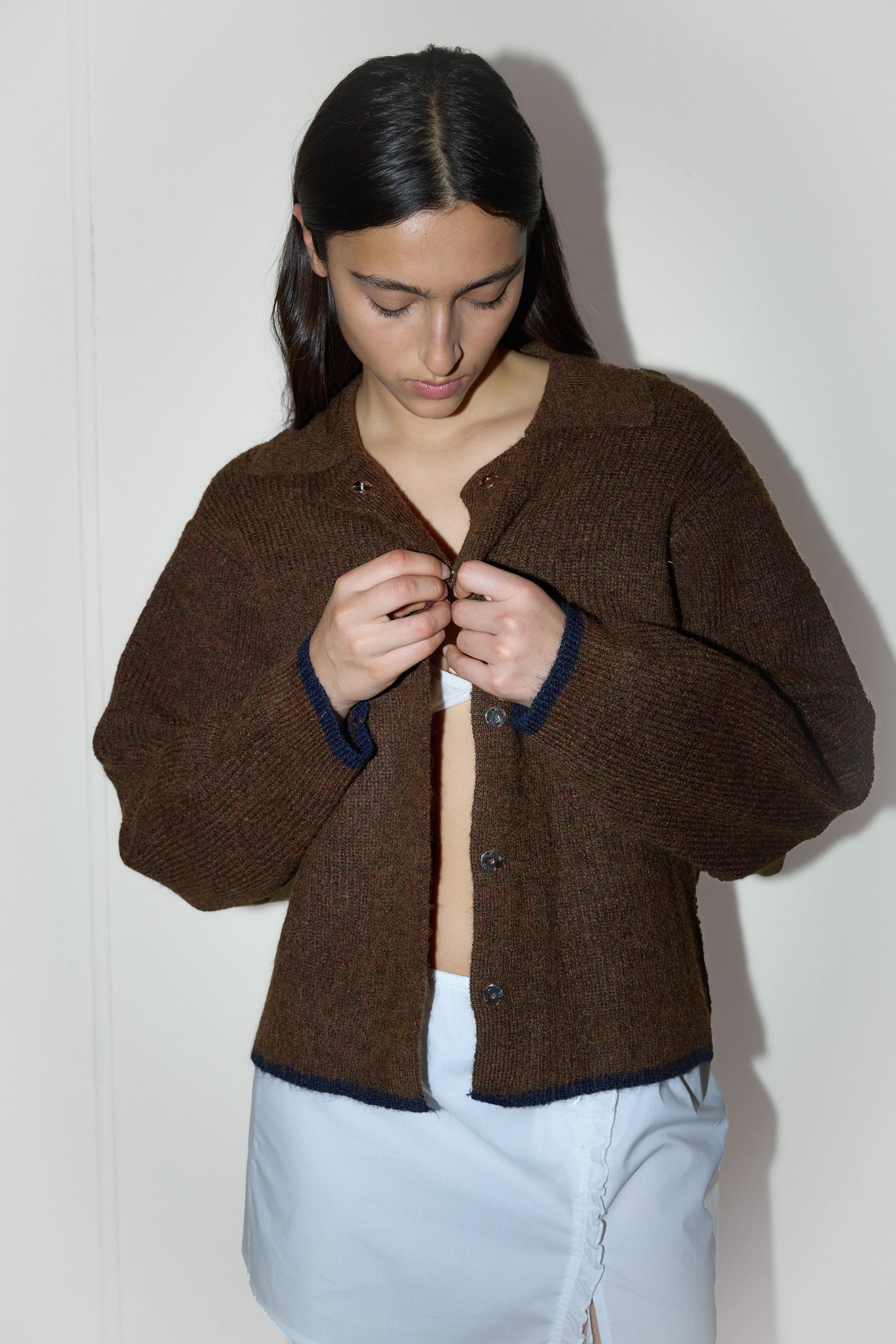 dark haired female model wears a brown elevated in a luxurious softly weighted and subtly lustrous sustainable knit blend featuring invisible press stud closure front, a relaxed collar and dropped shoulder, a gently curved arm for soft shaping and oversized fit for layering, paired with a white simple mini skirt designed to sit at low waist features adjustable tie and ruffle edge on side with button detail