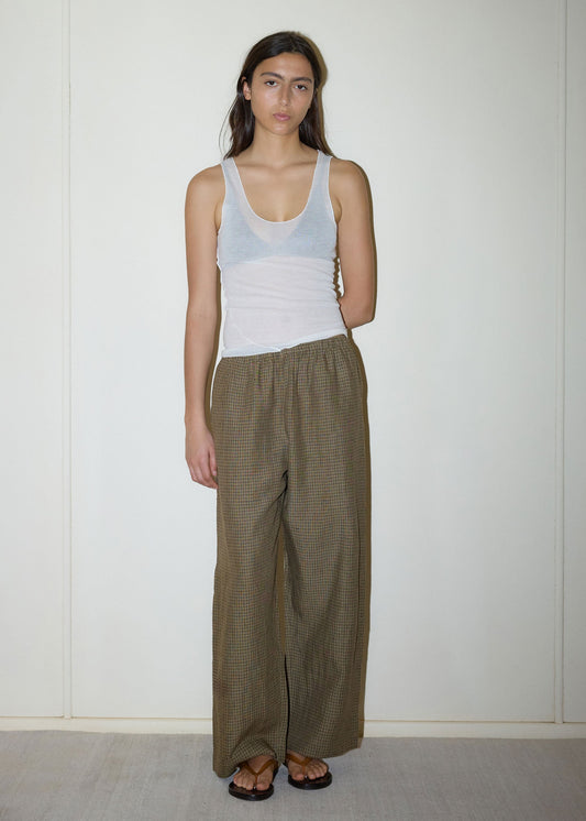 A person with long dark hair stands by a light wall, wearing a sheer white tank, Linen Ease Trouser - Pine Check by Deiji Studios, and dark sandals. One arm tucked behind them, they exude relaxed elegance in organic French linen.