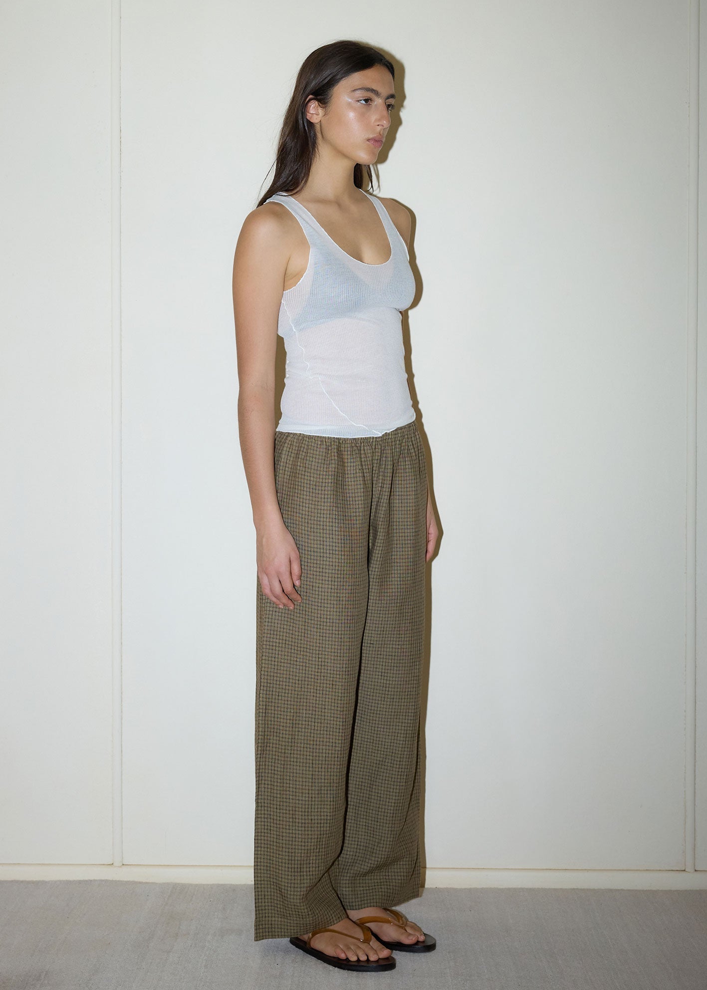 A person stands against a plain, light wall wearing a white sleeveless tank top, Deiji Studios' Linen Ease Trouser in Pine Check, and black flip-flops. They have long dark hair and a neutral expression, looking slightly to the side.