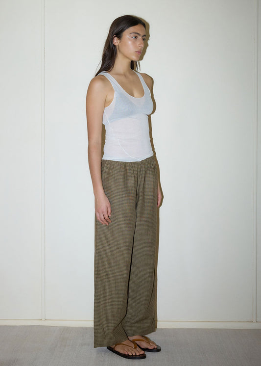 A woman stands indoors by a plain light wall, wearing a sheer white tank top, black flip-flops, and Ease Trouser—loose brown checkered pants. She faces left in a relaxed pose with her arms at her sides.