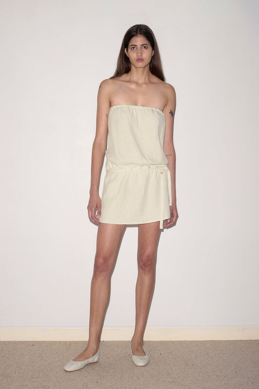 A woman with long brown hair wears the Strapless Linen Dress, a light beige mini dress with a gathered waist and flat beige shoes, standing against a plain white wall.