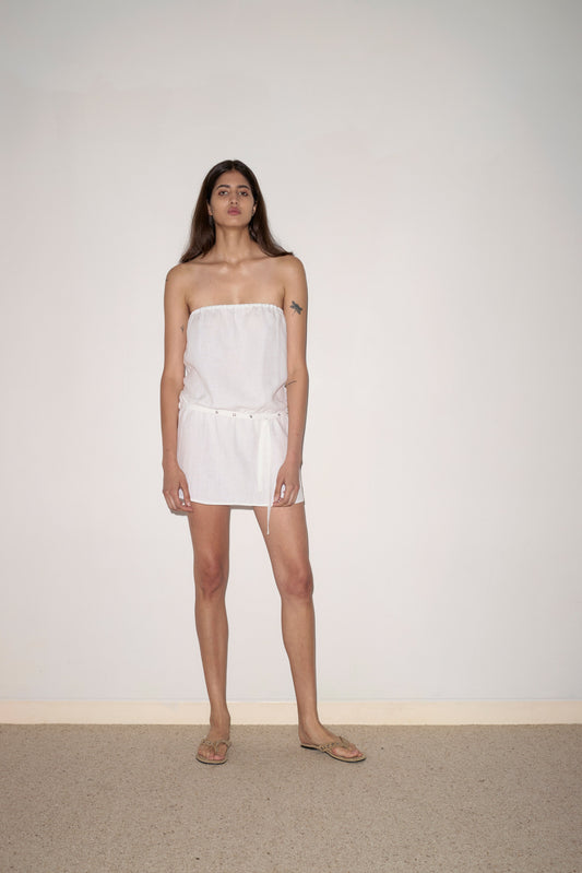 A person with long brown hair wears the Strapless Linen Dress, flip-flops, and a white belt, standing against a plain white wall and beige carpet. A small tattoo is visible on their upper left arm.