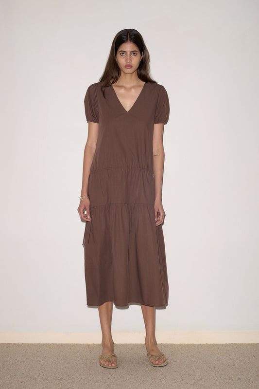 A woman with long hair and a neutral expression stands against a plain wall, wearing the Waistcord Dress - Carob Check by Deiji Studios, featuring short sleeves, a V-neck, tiered skirt details, and sandals for an effortless style.