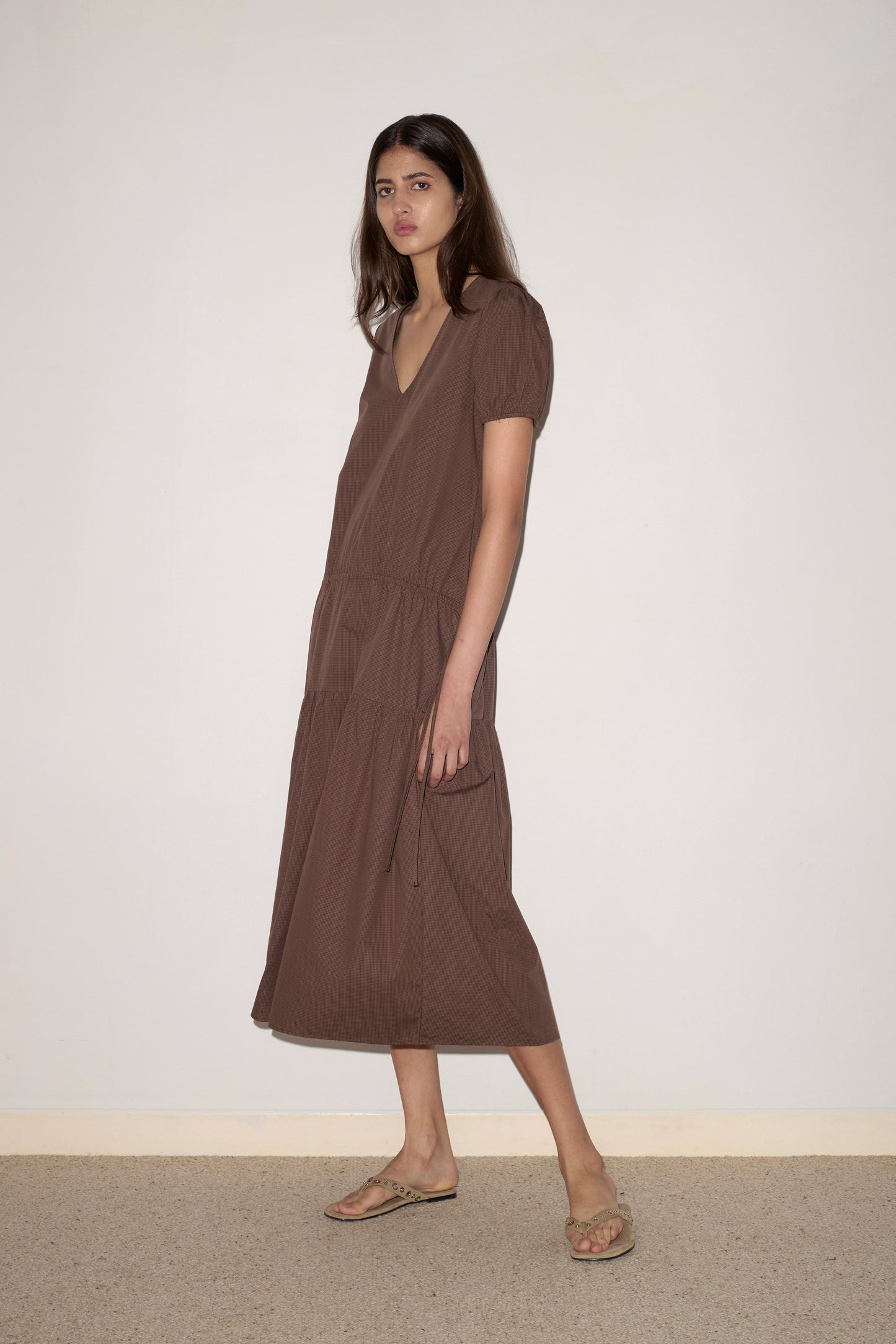 A woman with long brown hair wears the Waistcord Dress - Carob Check by Deiji Studios, a loose-fitting organic cotton dress in brown tones with short sleeves and a V-neck. She pairs it with flat sandals and stands on a beige carpet against a plain light wall.
