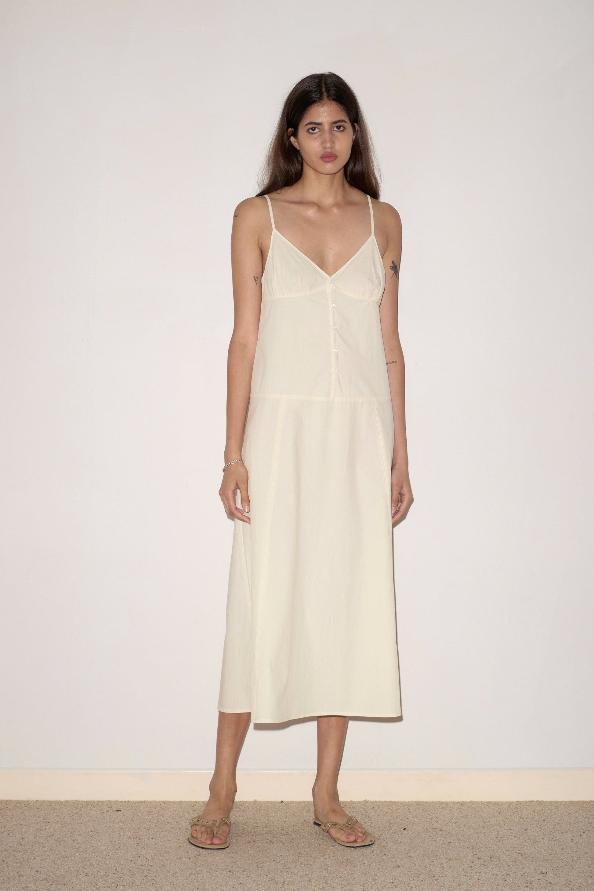 A woman with long brown hair stands against a plain, light wall, wearing the Double Seam Dress - Butter White by 0 and beige sandals. Her expression is neutral and her arms are relaxed by her sides.