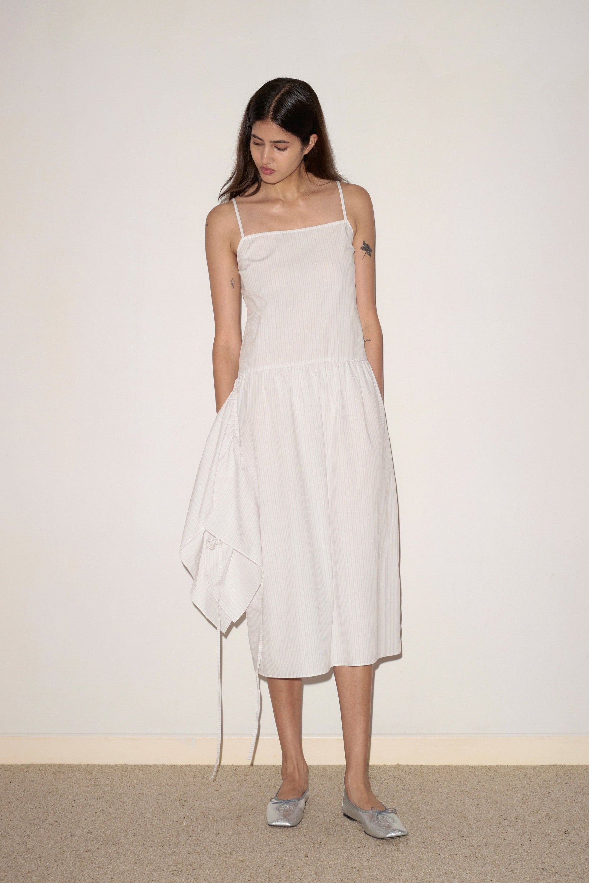 A woman with long brown hair wears the 0 Peplum Dress - Pacific Stripe, a sleeveless, knee-length style, as she stands indoors against a plain wall looking down with her hands by her sides.