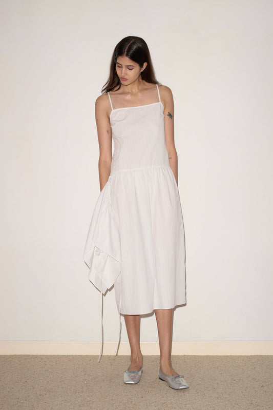 A woman with long brown hair wears the 0 Peplum Dress - Pacific Stripe, a sleeveless, knee-length style, as she stands indoors against a plain wall looking down with her hands by her sides.