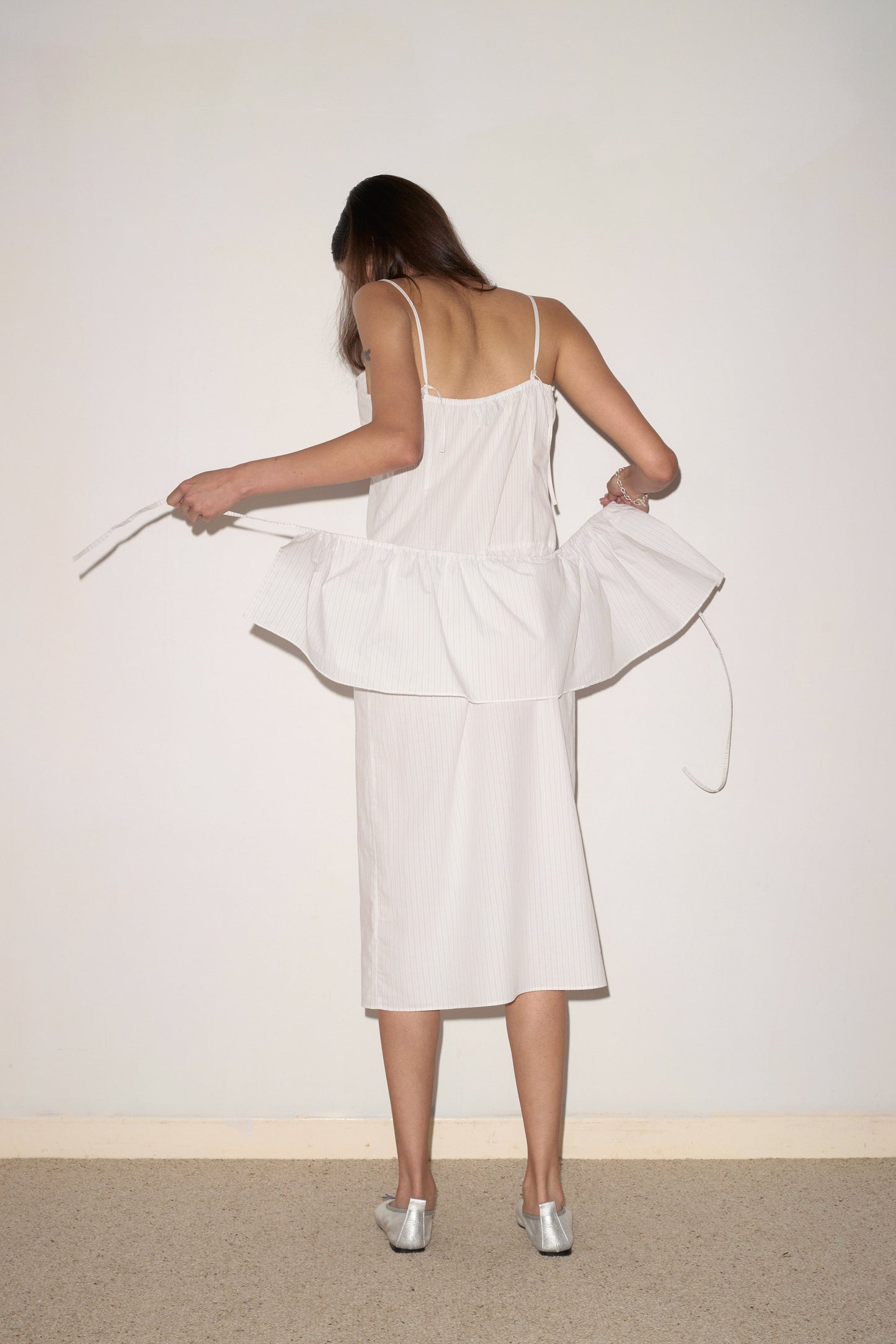 A woman with long brown hair, wearing the Peplum Dress - Pacific Stripe by 0 and silver shoes, stands facing a blank wall and adjusts the peplum detail. Her face is not visible.
