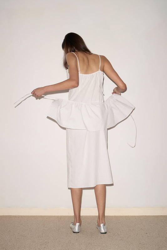 A woman with long brown hair, wearing the Peplum Dress - Pacific Stripe by 0 and silver shoes, stands facing a blank wall and adjusts the peplum detail. Her face is not visible.