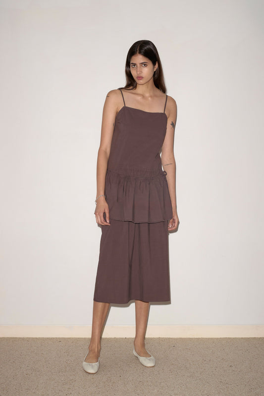 A woman with long brown hair stands against a light wall, wearing the Peplum Dress - Chocolate by 0. The loose, sleeveless dress features thin straps, a tiered skirt of organic cotton, and is paired with light flat shoes. She has a neutral expression.