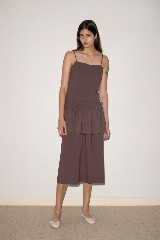 A woman with long brown hair stands against a light wall, wearing the Peplum Dress - Chocolate by 0. The loose, sleeveless dress features thin straps, a tiered skirt of organic cotton, and is paired with light flat shoes. She has a neutral expression.