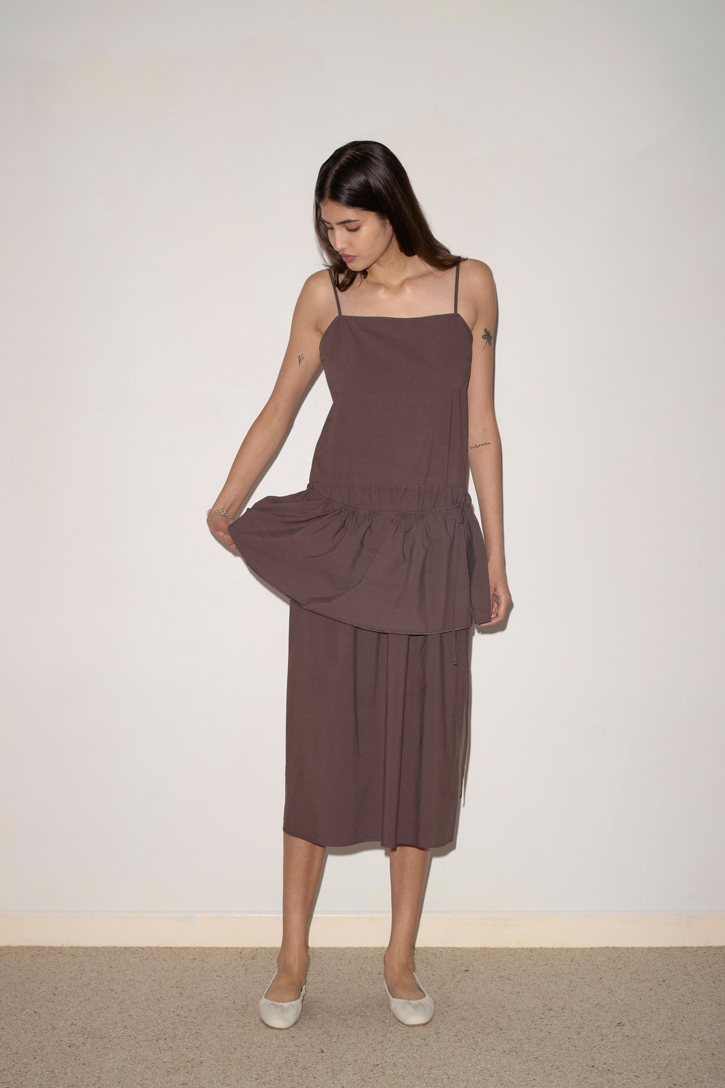 A woman wears the Peplum Dress - Chocolate by 0, standing indoors against a pale wall. She holds her skirt with one hand, looks down, and completes the look with white flat shoes.