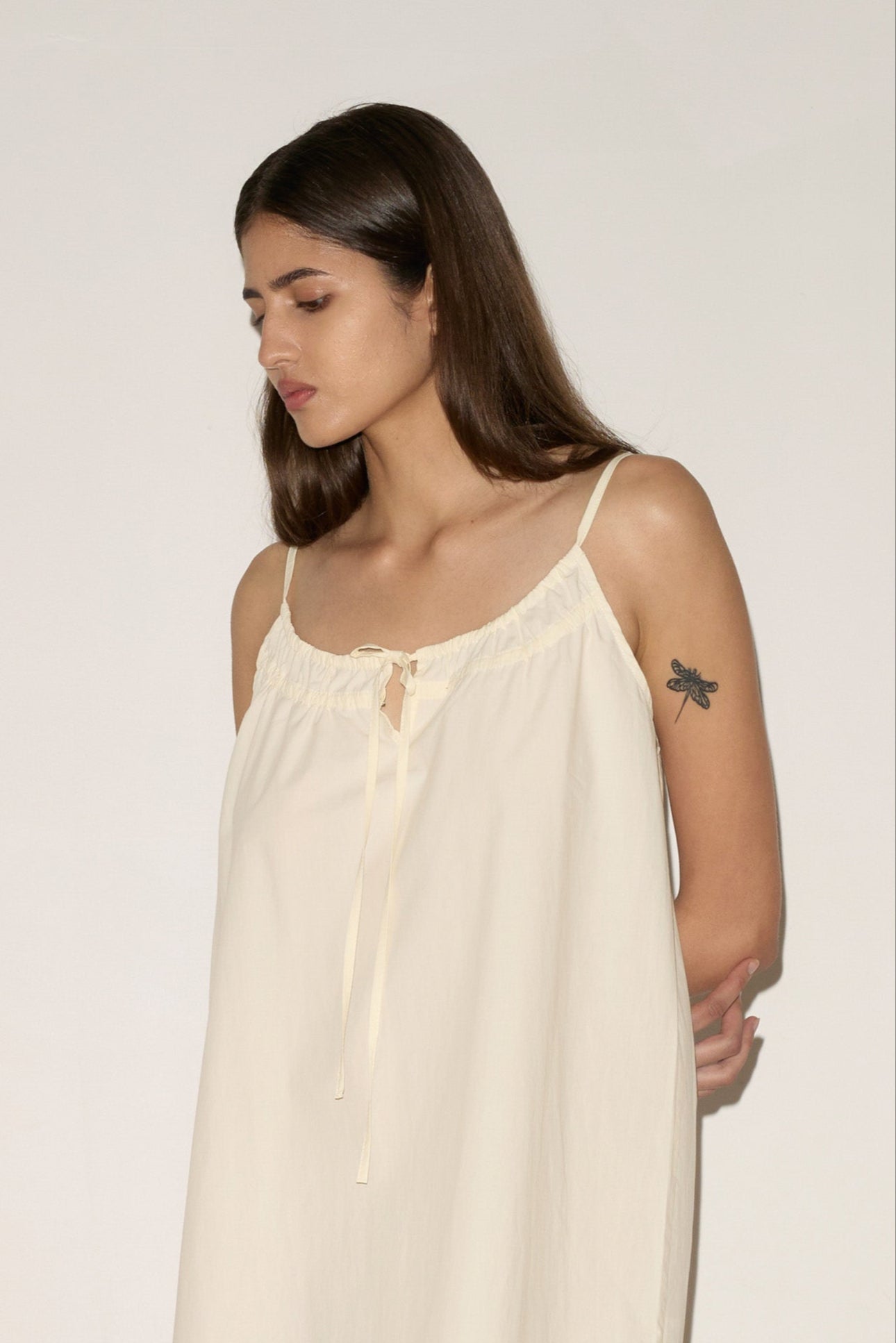 A woman with long brown hair stands indoors against a plain wall, wearing the Light Dress - Butter White by 0 and sandals. She has a tattoo on her upper left arm and her hands are behind her back.